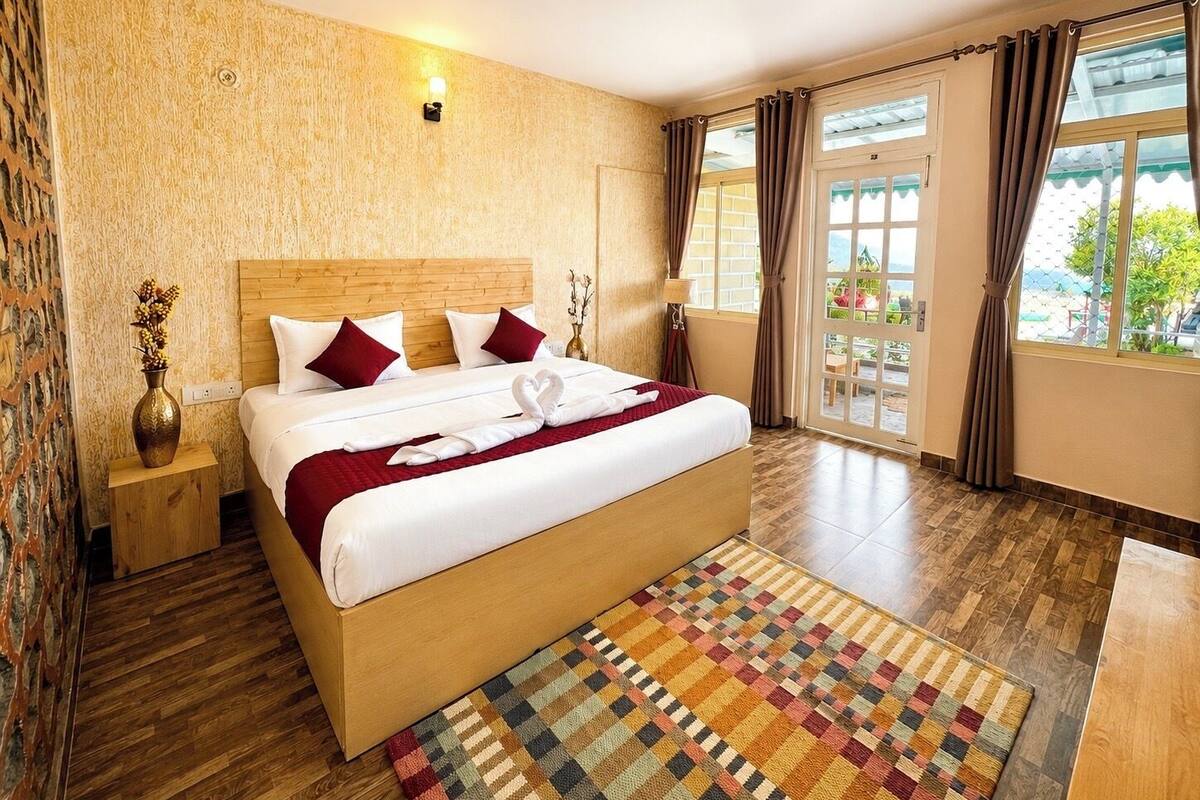 A hotel room with a wooden bed, a patterned rug, a vase with flowers, and a view of the outdoors through the window.