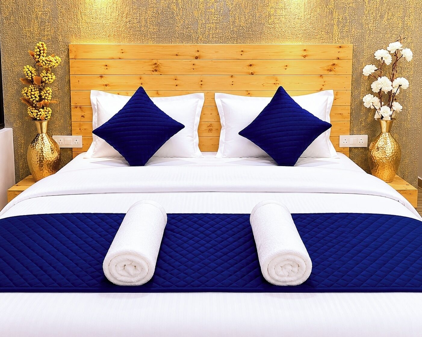 A neatly made bed with a wooden headboard, two blue pillows, and two rolled white towels.