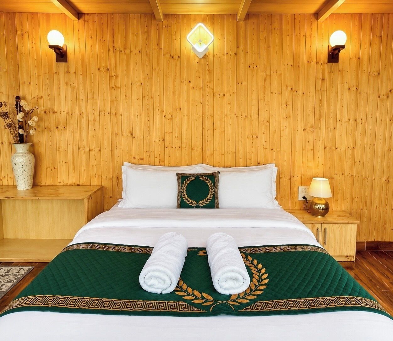 A wooden-paneled room with a bed, two wall sconces, a nightstand, and a vase with dried flowers.
