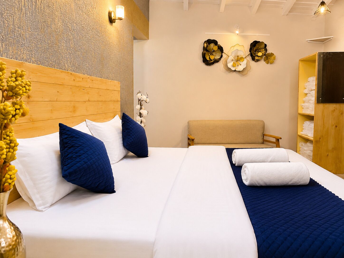 Habitación de hotel con cabecera de madera, dos camas, un banco, una lámpara fijada a la pared y un estante con toallas.