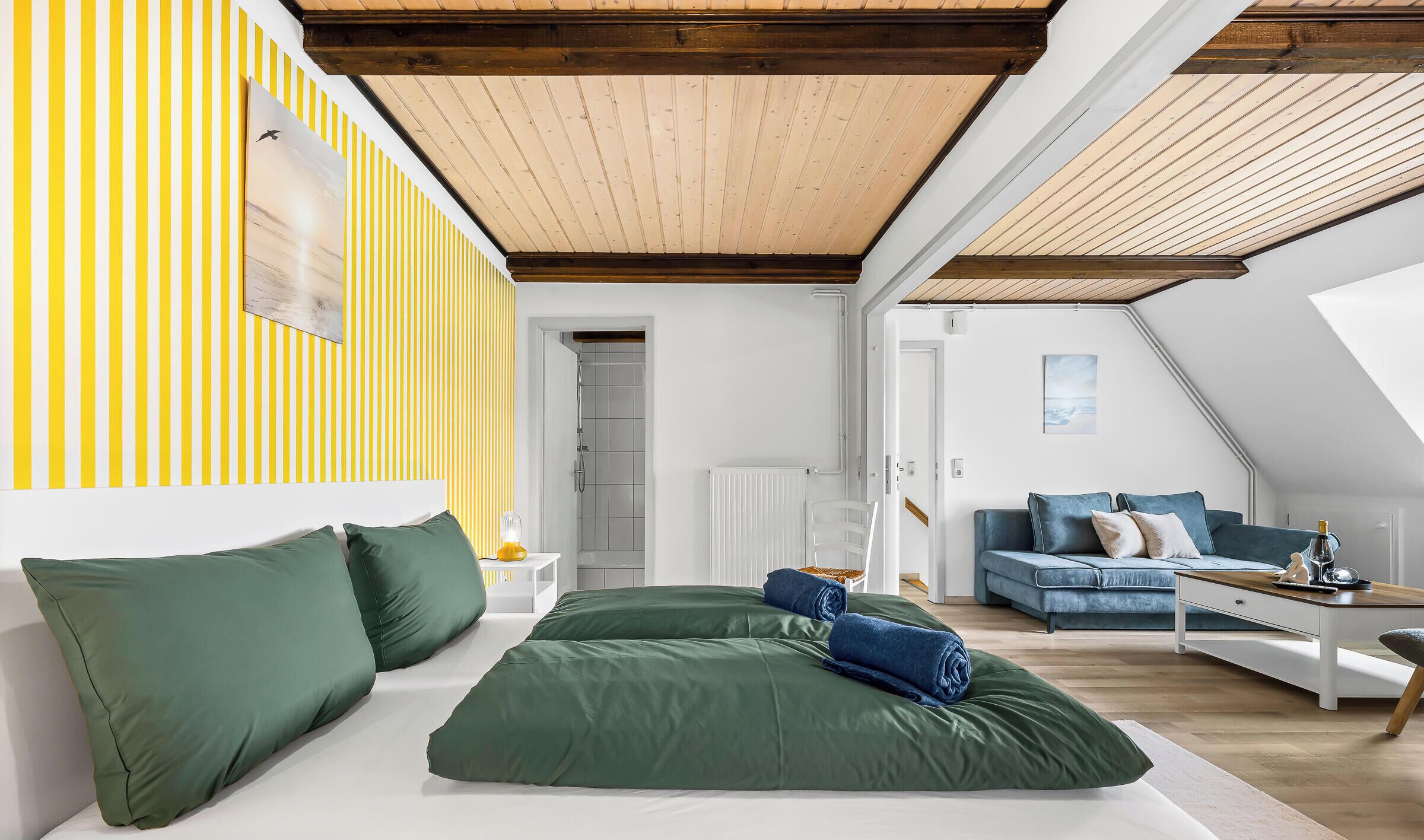 A modern loft bedroom with a sloped ceiling, a large bed with green bedding, a blue sofa, and a wooden floor.