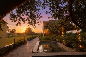 A serene outdoor setting with a pathway leading to a building, surrounded by lush greenery and a small water feature. - Achal Bagh – Boutique Stay Abu Road (Abu Road)