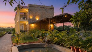 A hotel with a stone facade, a covered outdoor area with a thatched roof, and a landscaped garden with a water feature. - Achal Bagh – Boutique Stay Abu Road (Abu Road)