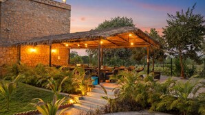 A stone building with a thatched roof patio area, surrounded by lush greenery and outdoor seating. - Achal Bagh – Boutique Stay Abu Road (Abu Road)