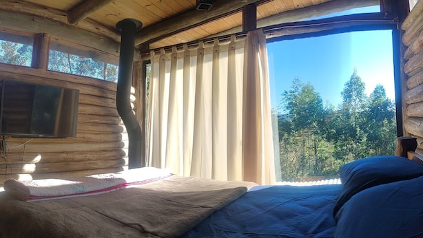 Une chambre dans un chalet en rondins, avec un lit, un téléviseur fixé au mur et une fenêtre avec des rideaux.
