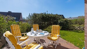 Eine Terrasse mit gelb-weiß gestreiften Stühlen und einem weißen Tisch, umgeben von Grünpflanzen und einer Backsteinmauer.