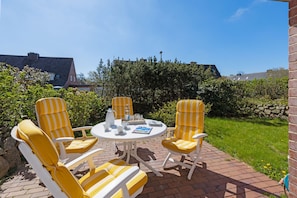 Eine Terrasse mit gelb-weiß gestreiften Stühlen und einem weißen Tisch, umgeben von Grünpflanzen und einer Backsteinmauer.