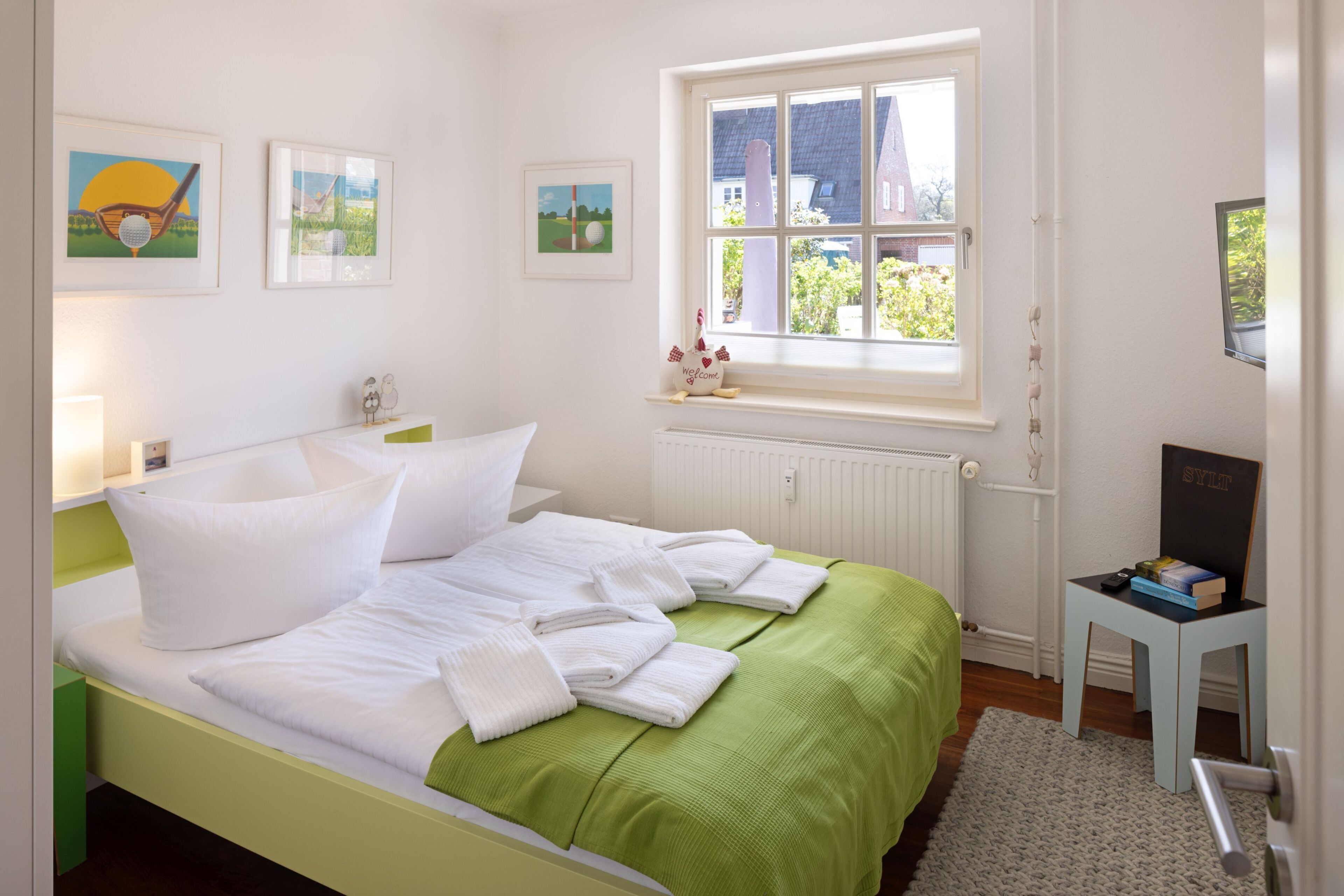 A neatly made bed with white linens and a green bedspread, a window with a view of a house, and a small table with a book and a chalkboard.