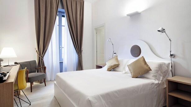 A hotel room with a large bed, a desk with a lamp, a chair, and a view of the outdoors through a window with curtains. - Guesthouse Delle Navi - Room 4 (Roma)