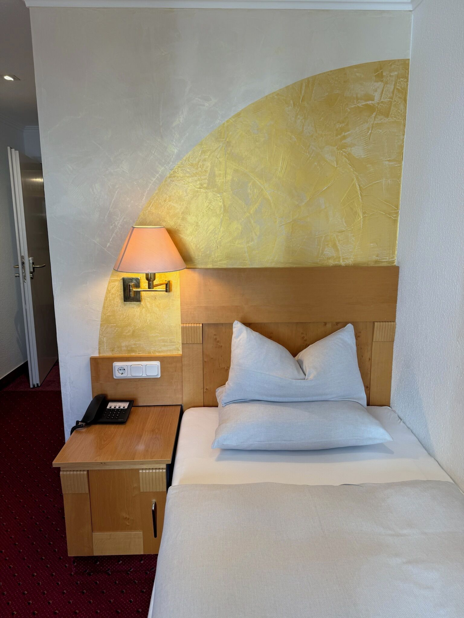 A hotel room with a bed, a bedside table with a lamp, a phone, and a wall-mounted light fixture.