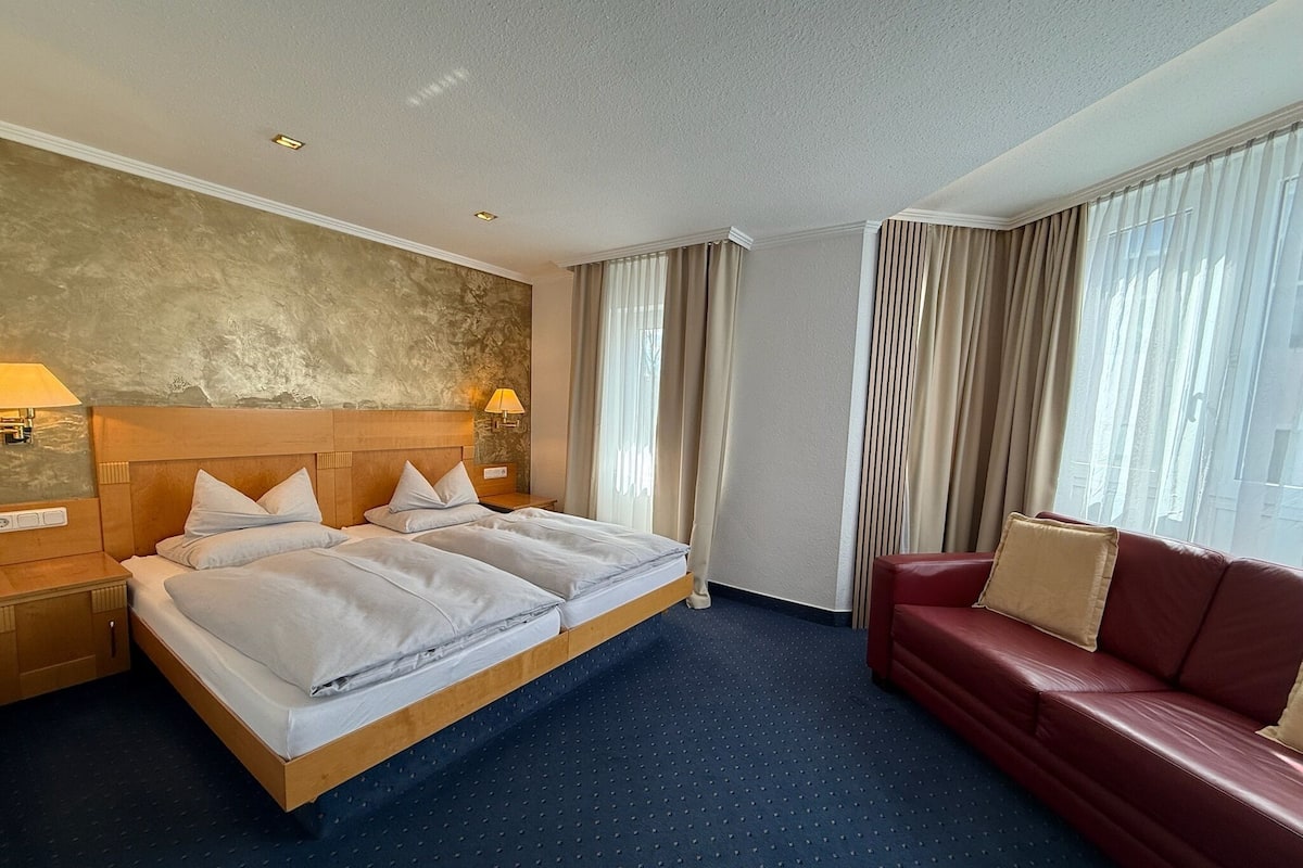 Habitación de hotel con una cama grande, un sofá rojo, una lámpara de pared y una ventana con cortinas.