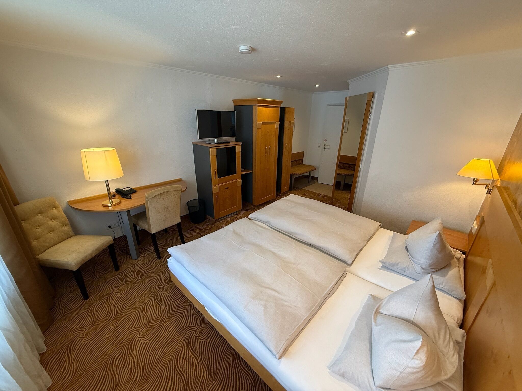 Una habitación de hotel con una cama, un escritorio con lámpara, una silla, un armario y una televisión.