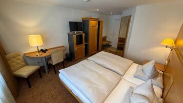 Quarto de hotel com cama, escrivaninha com luminária, cadeira, guarda-roupa e televisão.