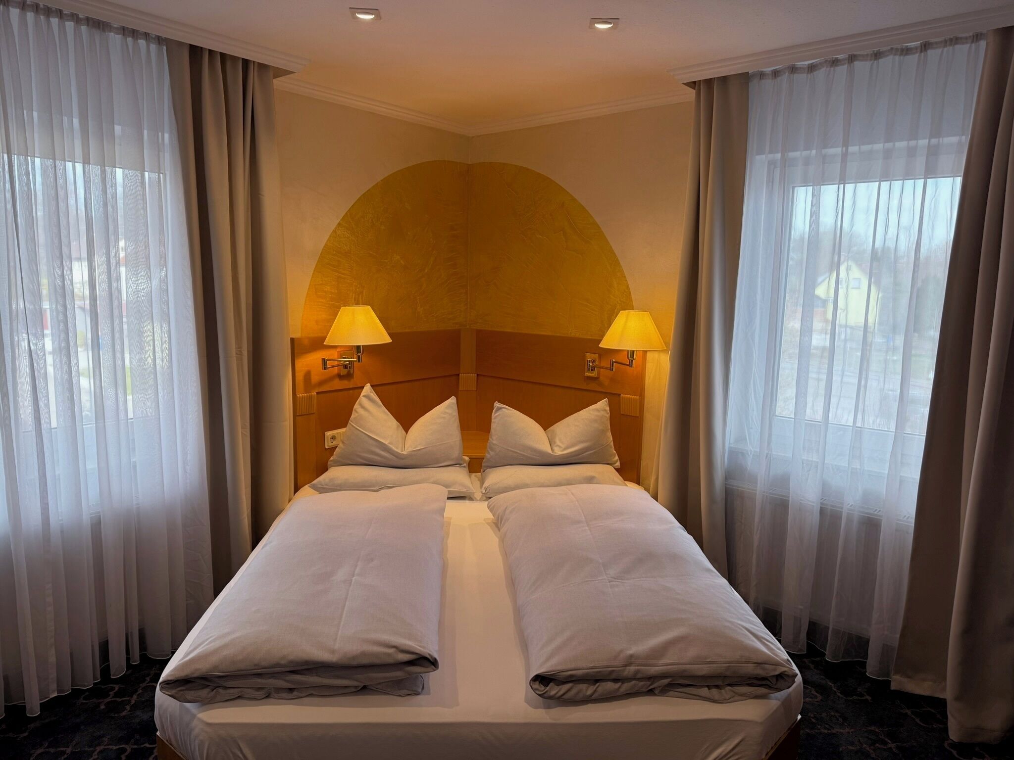 A double bed with two pillows, a headboard, two wall-mounted lamps, and sheer curtains.
