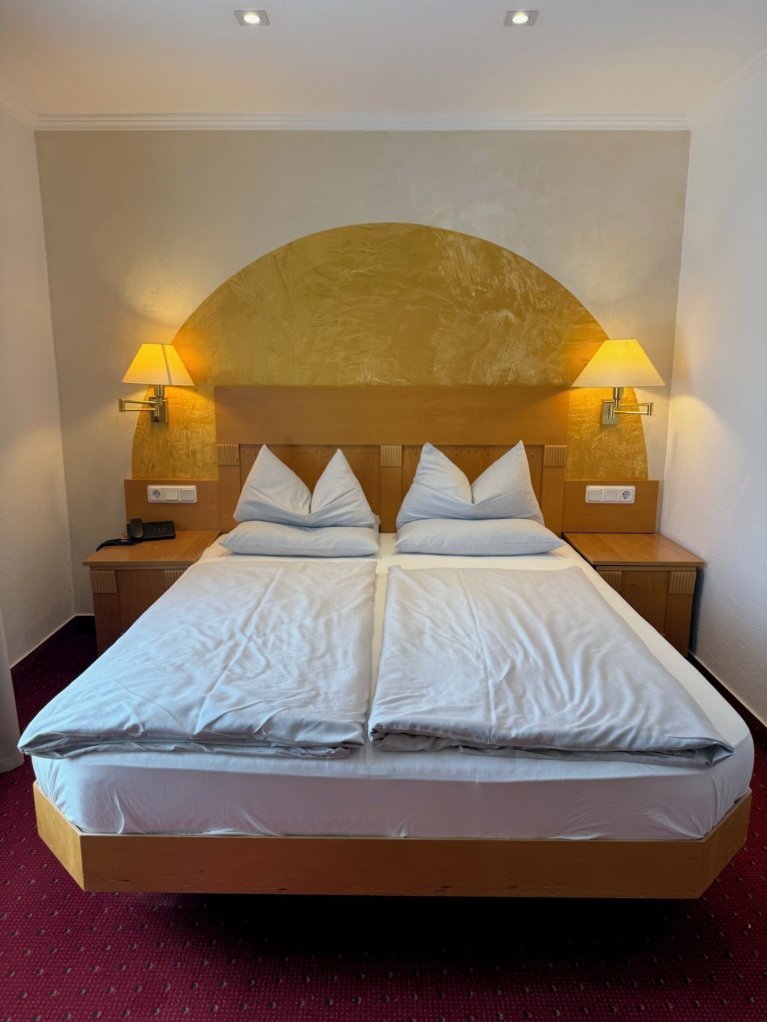 Un lit double avec une tête de lit en bois, deux tables de chevet avec des lampes et un sol recouvert d’une moquette rouge.