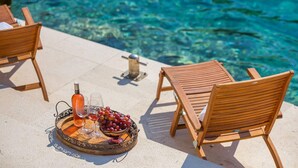 A poolside setting with wooden lounge chairs, a tray with a bottle, glasses, and grapes. - Seafront Villa Marella (Milna)