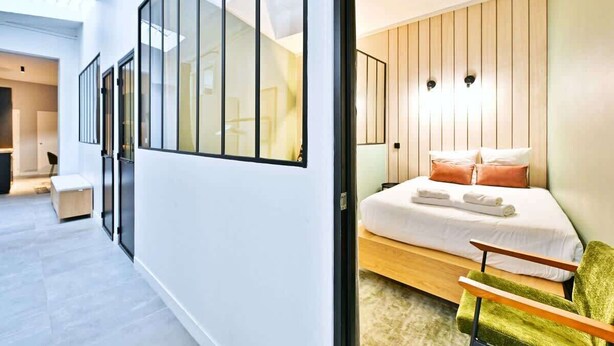 A modern hotel room with a large bed, a green chair, and a view of the city through floor-to-ceiling windows. - Loft moderne 2BR - Cœur de Paris A/C portable (Paris)