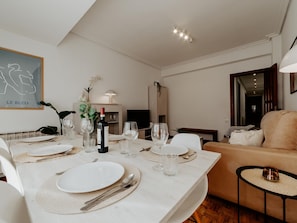 Et spiseområde med et bord dækket til fire personer, en sofa og et fjernsyn i baggrunden.