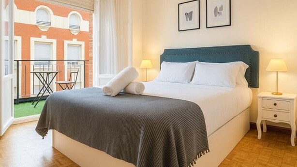 Une chambre d’hôtel comprenant un lit, deux lampes de chevet, une fenêtre avec des rideaux et un balcon avec une table et des chaises.