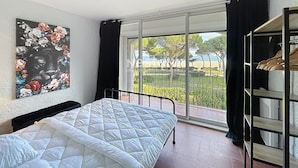 A bedroom with a bed, a large window with a view of trees and a beach, and a black curtain.