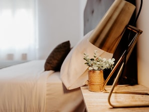 Ein ordentlich bezogenes Bett mit Kopfteil, ein Nachttisch mit Pflanze und eine Lampe.
