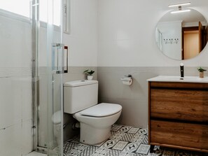 Ein Badezimmer mit Toilette, Waschbecken und Spiegel. Ein Duschbereich mit Glastür. Der Boden ist mit gemusterten Fliesen ausgelegt.