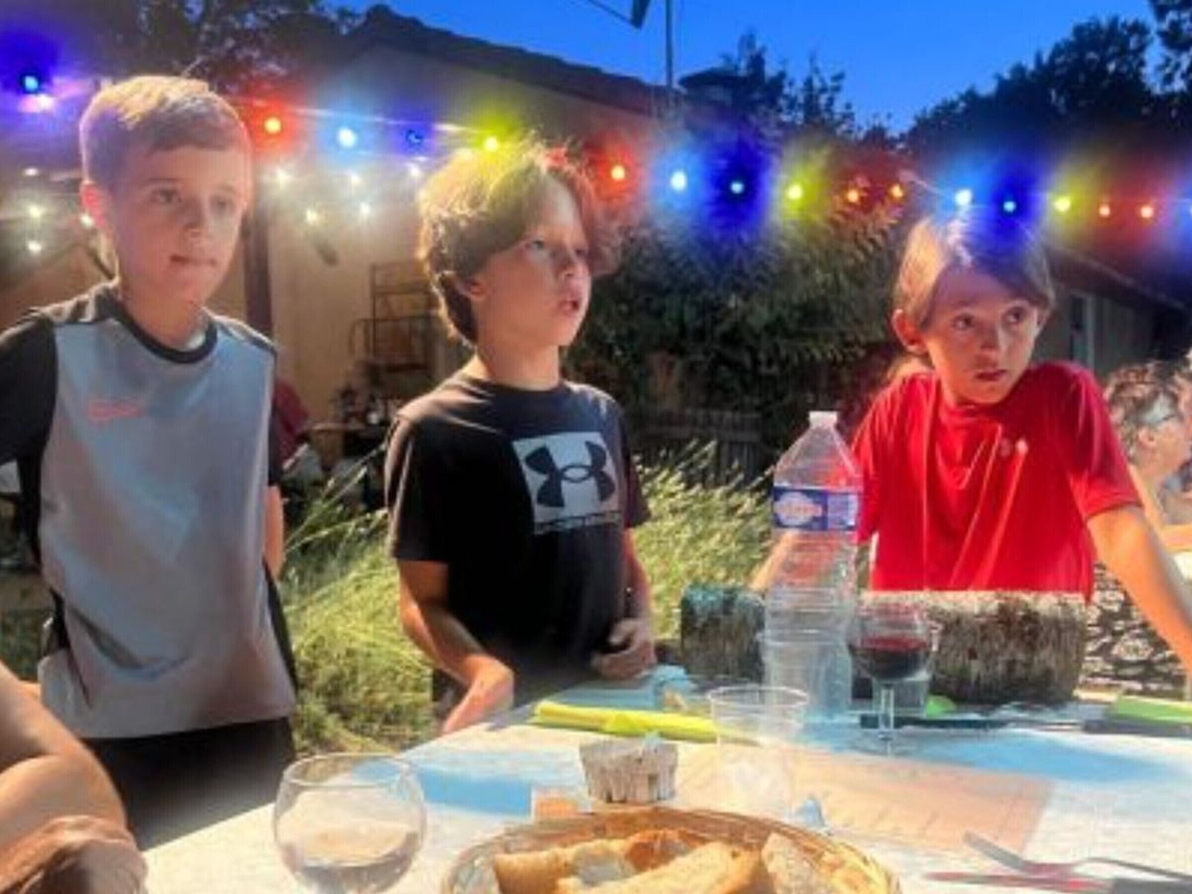 Une table avec des aliments et une bouteille d’eau, installée à l’extérieur, avec des guirlandes lumineuses en arrière-plan.