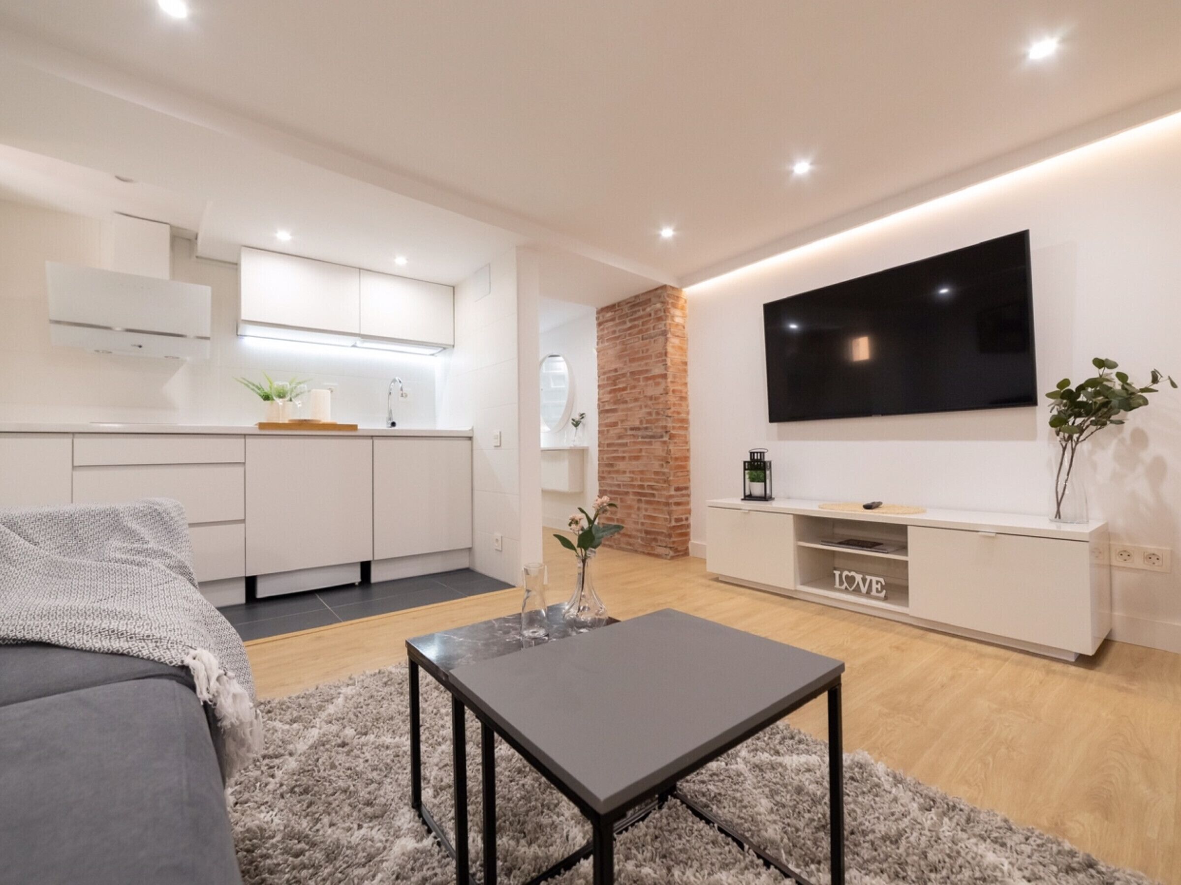 Un salón moderno con un sofá gris, una mesita, un televisor montado en la pared y una cocina con armarios blancos y una pared de ladrillo decorativa.