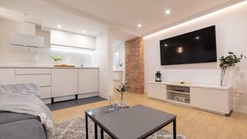 Un salón moderno con un sofá gris, una mesita, un televisor montado en la pared y una cocina con armarios blancos y una pared de ladrillo decorativa.