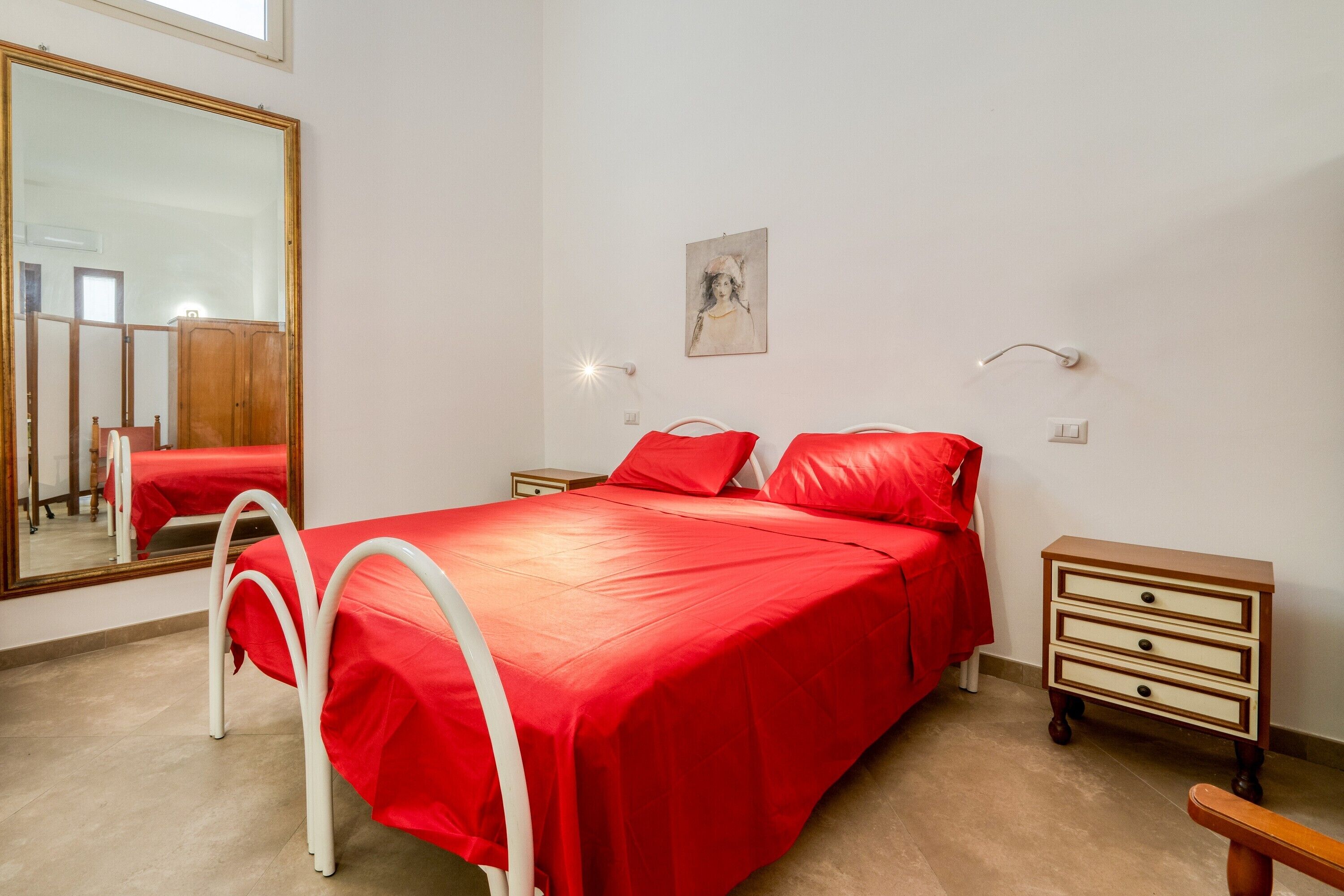 Un dormitorio con cama, mesita de noche, cómoda, espejo y lámpara de pared.