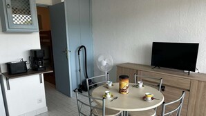 Un espacio habitable compacto con kitchenette, una zona de comedor con cuatro sillas, cafetera, ventilador y televisor.