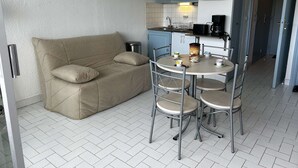 Un espacio habitable compacto con un sofá, una pequeña mesa de comedor con sillas, una zona de cocina con armarios y electrodomésticos, y una puerta que da a otra habitación.