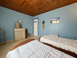 A bedroom with two beds, a wooden ceiling, a door, a chair, a small cabinet, and a surfboard on the wall. - House with Garden in Saint-Georges-d'Oléron for 6 people (Saint-Georges-d'Oléron)