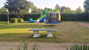 A park with a green and blue inflatable slide, a picnic table, and a hedge in the foreground. - Mobile Home ADA 28m², 4 pers, Terrace (Marcilly-sur-Vienne)
