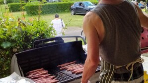 A person grilling hot dogs outdoors with a view of a manicured garden and parked cars in the background. - Mobile Home ADA 28m², 4 pers, Terrace (Marcilly-sur-Vienne)
