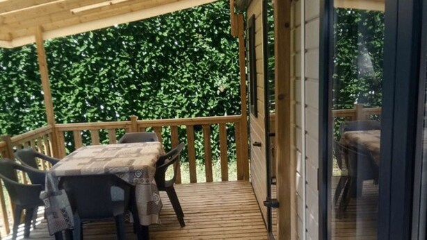 A covered outdoor area with a wooden deck, a table with chairs, and a view of greenery in the background. - Family Mobile Home with Terrace - 4 People (Marcilly-sur-Vienne)