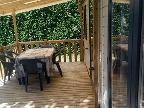 A covered outdoor area with a wooden deck, a table with chairs, and a view of greenery in the background. - Family Mobile Home with Terrace - 4 People (Marcilly-sur-Vienne)