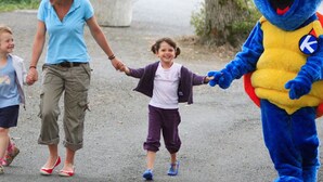 Une personne déguisée en dinosaure bleu, tenant la main de deux enfants qui marchent sur un sentier.