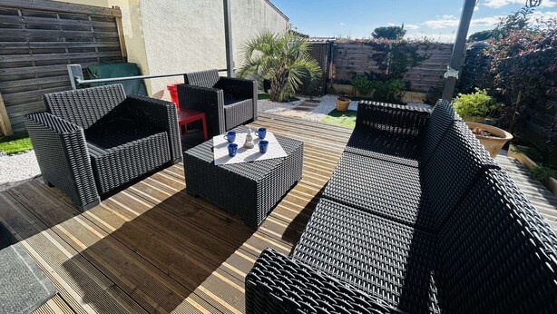 Eine Terrassengruppe im Freien mit Korbmöbeln, einem Tisch mit Tassen und Blick auf Garten und Haus.