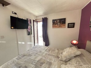 A bedroom with a bed, a flat-screen TV mounted on the wall, a window with curtains, and a wall-mounted lamp. - Cap d'Agde - T2 Apt with Garden, Saint Martin (Cap d'Agde)