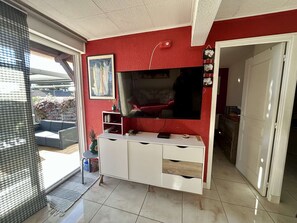 A living room with a red wall, a flat-screen TV, a white entertainment unit, and a sliding glass door leading to a balcony. - Cap d'Agde - T2 Apt with Garden, Saint Martin (Cap d'Agde)