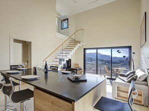 Eine moderne Küche mit Kochinsel, ein Essbereich mit Barhocker, eine Treppe mit Glastrennwand und ein Balkon mit Bergblick.