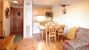 Een kleine keuken met houten kastjes, een magnetron en een eethoek met een tafel en stoelen.