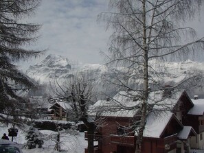 Ein verschneites Dorf mit traditionellen Holzhäusern, ein Berg im Hintergrund und kahl stehende Bäume.