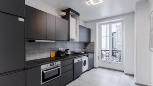 A modern kitchen with dark cabinetry, a built-in oven, a washing machine, and a window with a balcony. - Elegant 3BR Bastille Apartment (Paris)