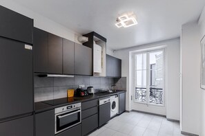 A modern kitchen with dark cabinetry, a built-in oven, a washing machine, and a window with a balcony. - Elegant 3BR Bastille Apartment (Paris)