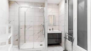 A modern bathroom with a glass-enclosed shower, a vanity with a mirror, and a large window. - Elegant 3BR Bastille Apartment (Paris)