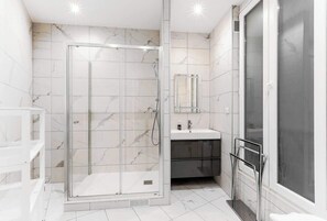 A modern bathroom with a glass-enclosed shower, a vanity with a mirror, and a large window. - Elegant 3BR Bastille Apartment (Paris)