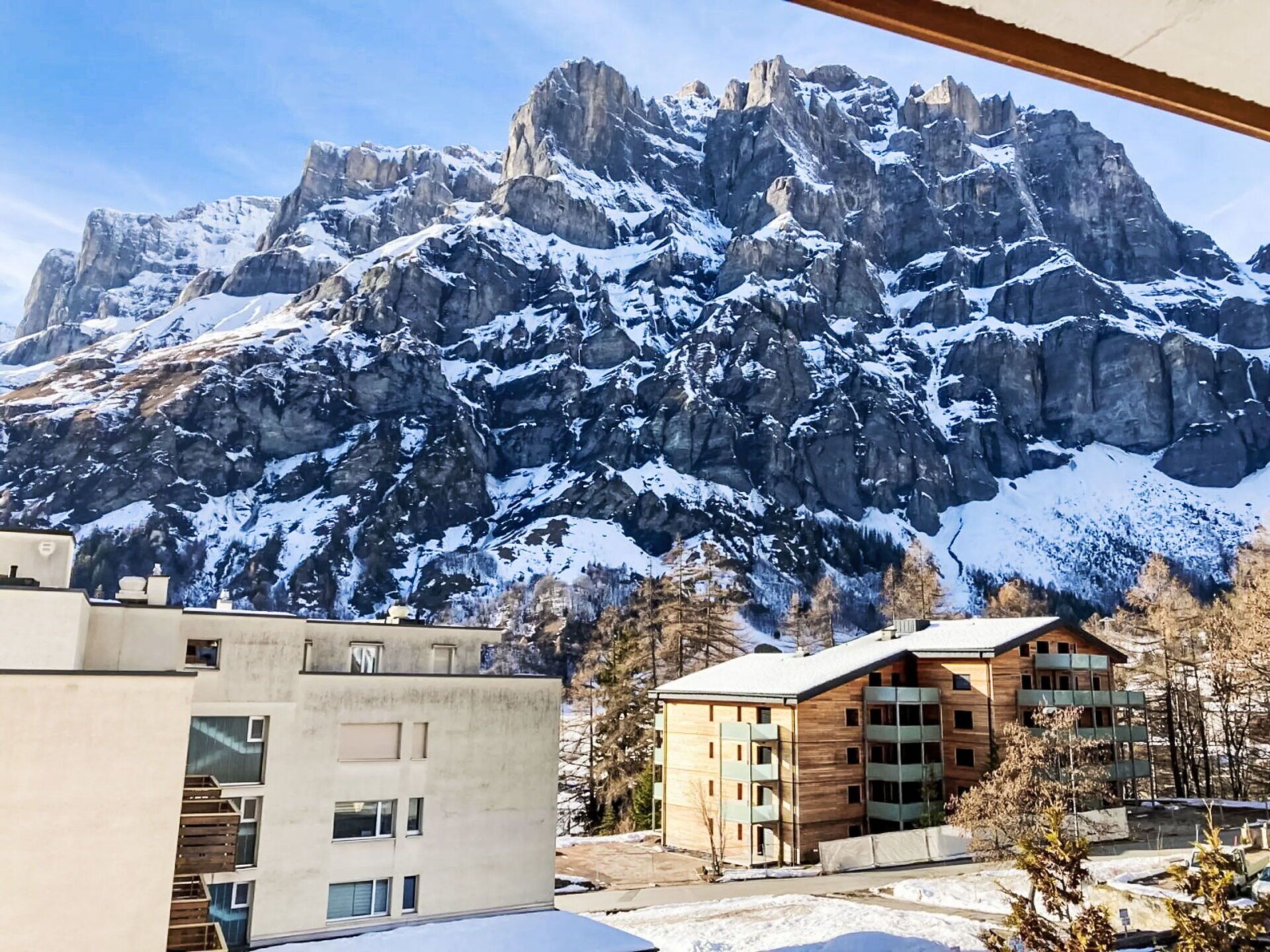 A modern building with a wooden facade and multiple balconies, situated in a snowy landscape with a mountain backdrop.