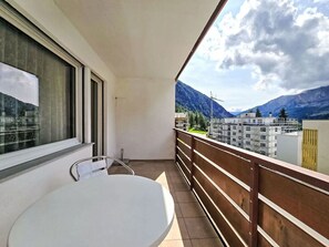 En balkon med et hvidt bord og en stol, et trægelænder og udsigt til bjerge og bygninger.